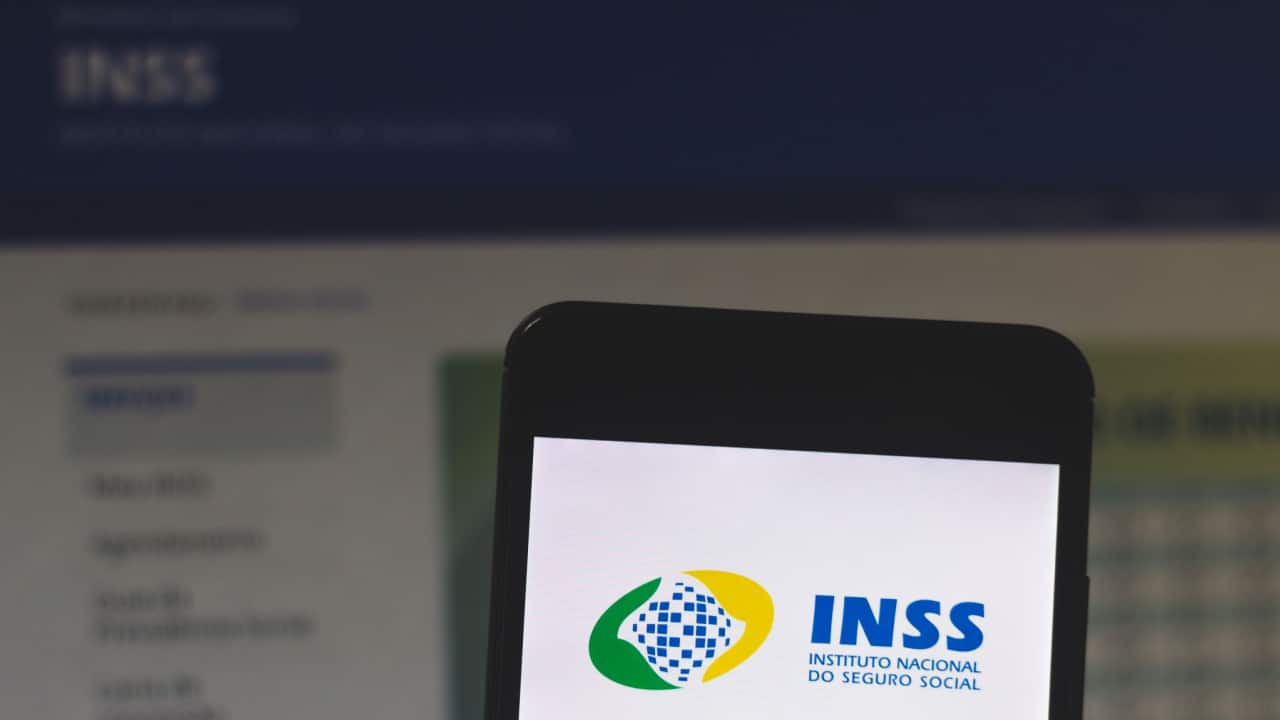 Ao fundo, site do INSS aberto e celular na frente com logo do INSS