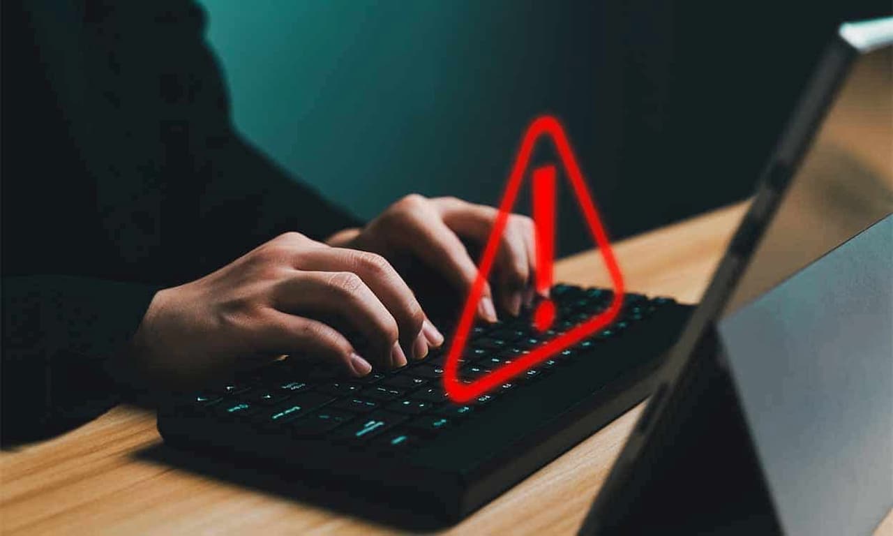 Pessoa digitando em um teclado de computador. Em cima, um sinal de alerta em vermelho. golpes