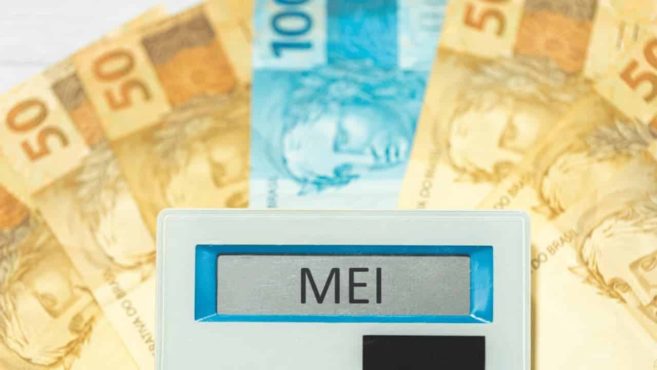 Calculadora com a palavra 'MEI' escrita em seu visor e cédulas de 100 e 50 reais atrás. governo