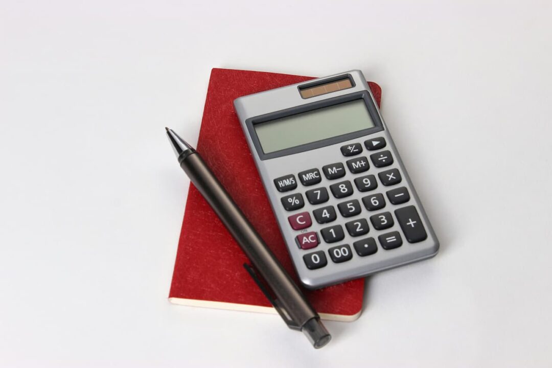 Imagem de uma calculadora e uma caneta em cima de um caderno em um fundo branco
